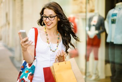 Eine Frau mit mehreren Einkaufstaschen schaut glücklich auf ihr Mobiltelefon.