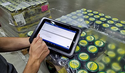 virtuelle Unterschrift auf dem Tablet bei Warenübergabe, im Hintergrund Palette mit Gurkengläsern