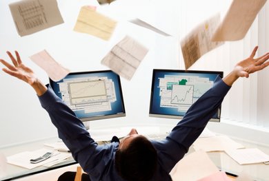 Ein Mann sitzt an einem Schreibtisch vor zwei Monitoren, auf denen verschiedene Grafiken zu sehen sind. Er wirft zahlreiche Unterlagen in die Luft.