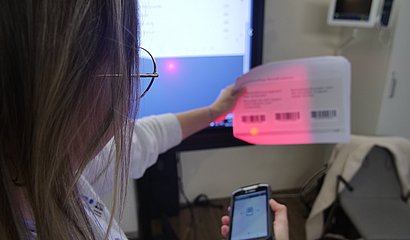 Ein Frau mit dem Rücken zur Kamera gewandt scannt im Krankenhauszimmer ein Blatt Papier mit Barcodes