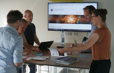 Foto: Gruppe steht an einem Tisch, im Hintergrund ein Screen mit der Darstellung einer Blockchain