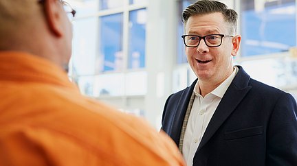 Mann in Anzug diskutiert angeregt und kooperativ mit einem Mann in orangefarbenem Hemd