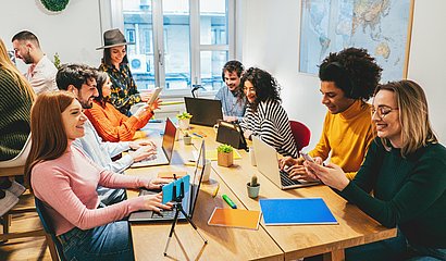 Junge stylische Leute sitzen gemeinsam im lichtdurchfluteten Raum am großen Tisch und nutzen verschiedene Devices wie Notebooks und Handys