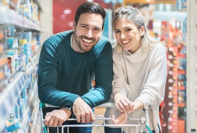 Eine Frau und ein Mann schieben lachend einen Einkaufswagen neben gefüllten Supermarktregalen.