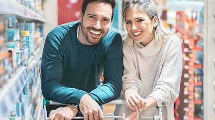 Eine Frau und ein Mann schieben lachend einen Einkaufswagen neben gefüllten Supermarktregalen.