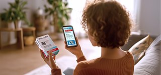 Eine Frau mittleren Alters sitzt im Wohnzimmer und scannt mit ihrem Smartphone einen GS1 DataMatrix Code auf einer Arzneimittelpackung