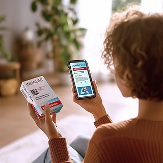 Eine Frau mittleren Alters sitzt im Wohnzimmer und scannt mit ihrem Smartphone einen GS1 DataMatrix Code auf einer Arzneimittelpackung