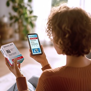 Eine Frau mittleren Alters sitzt im Wohnzimmer und scannt mit ihrem Smartphone einen GS1 DataMatrix Code auf einer Arzneimittelpackung