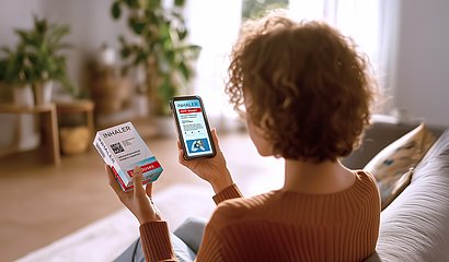 Eine Frau mittleren Alters sitzt im Wohnzimmer und scannt mit ihrem Smartphone einen GS1 DataMatrix Code auf einer Arzneimittelpackung