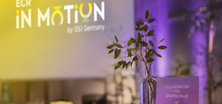 Tisch im Vordergrund mit Pflanze im Glas und einem Kärtchen, auf dem das Motto von ECR iN MOTiON „Collaboration for Shopper Value“ steht, und der Hintergrund ist in Gelb und Flieder gehalten
