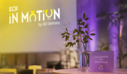 Premiere ECR iN MOTiON 2026 Tisch im Vordergrund mit Pflanze im Glas und einem Kärtchen, auf dem das Motto von ECR iN MOTiON „Collaboration for Shopper Value“ steht, und der Hintergrund ist in Gelb und Flieder gehalten