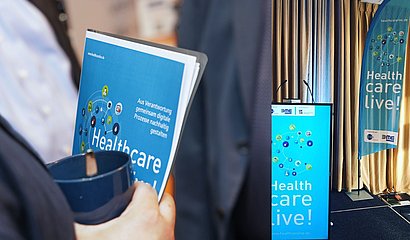 Fotokollage aus Programmflyer tragende Person und Healthcare live gebrandetes Rednerpult 