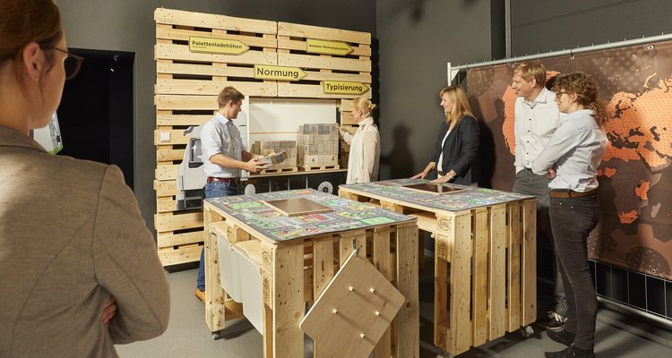 Technologies Experience - Eine Gruppe von Personen schaut zu, wie zwei Personen kleine Kisten auf Paletten legen