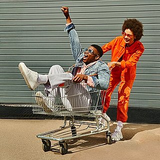 Eine junge Frau schiebt lachend einen jungen Mann in einem Einkaufswagen