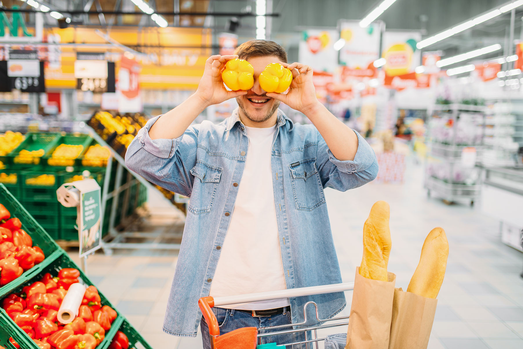Ein junger Mann im Supermarkt hält sich zwei gelbe Paprikas vor die Augen